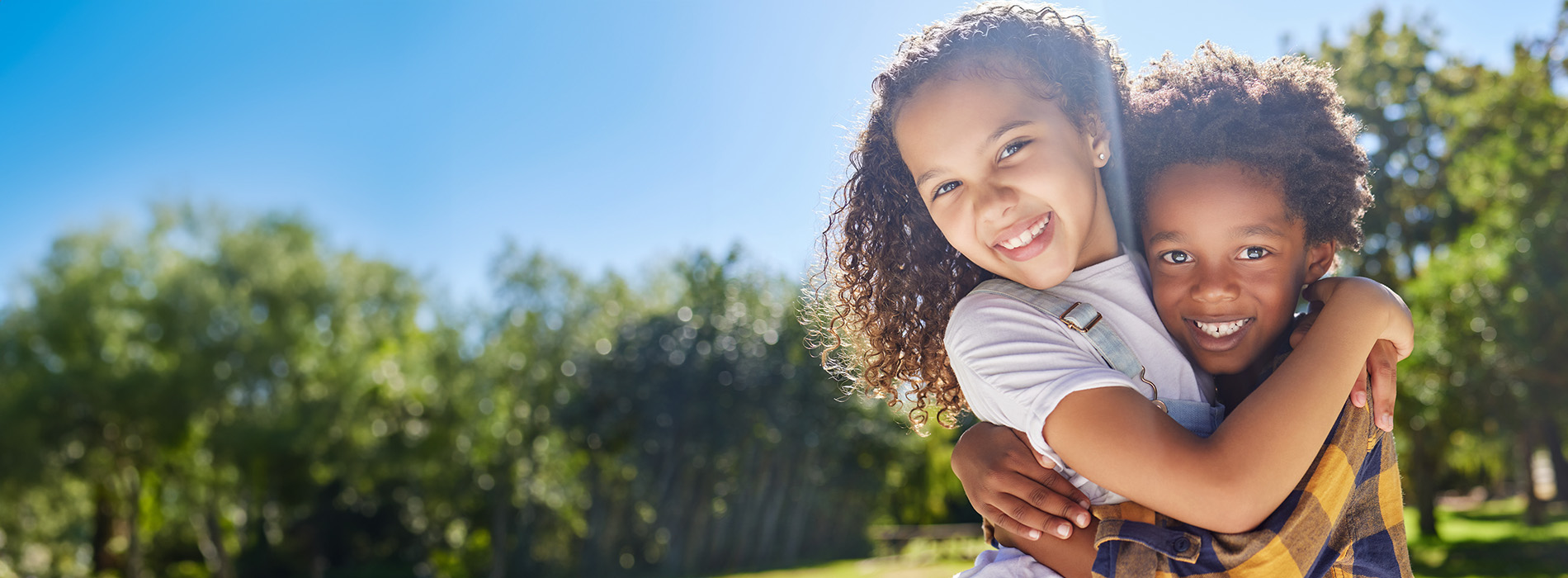 A young girl hugging an older boy with a clear blue sky in the background.
